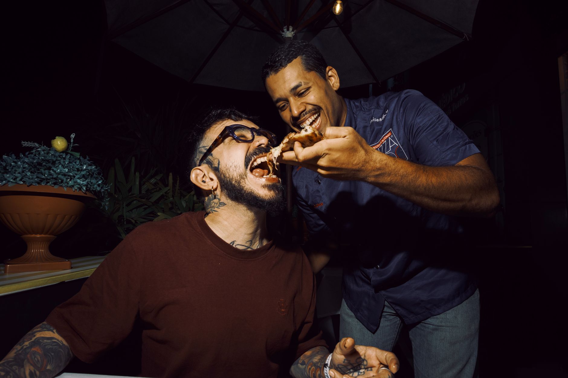 Two friends sharing a slice of pizza under a patio umbrella at night
